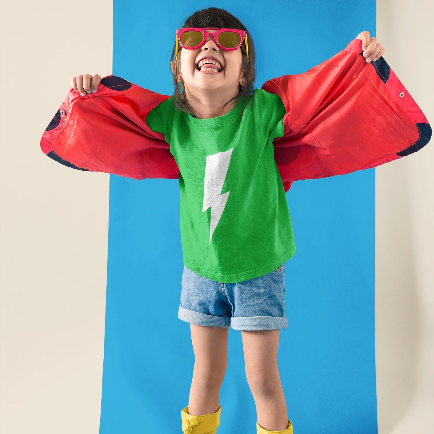 Child in superhero costume with red cape, green shirt with white lightning bolt, denim shorts, and yellow boots on a blue background.