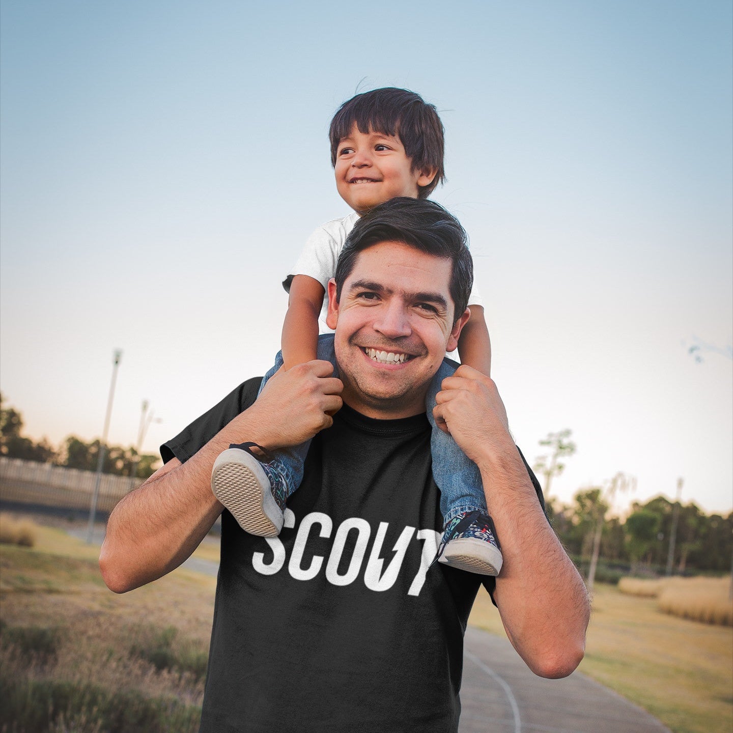 Man carrying a child on his shoulders with a Scout shirt, outdoors.